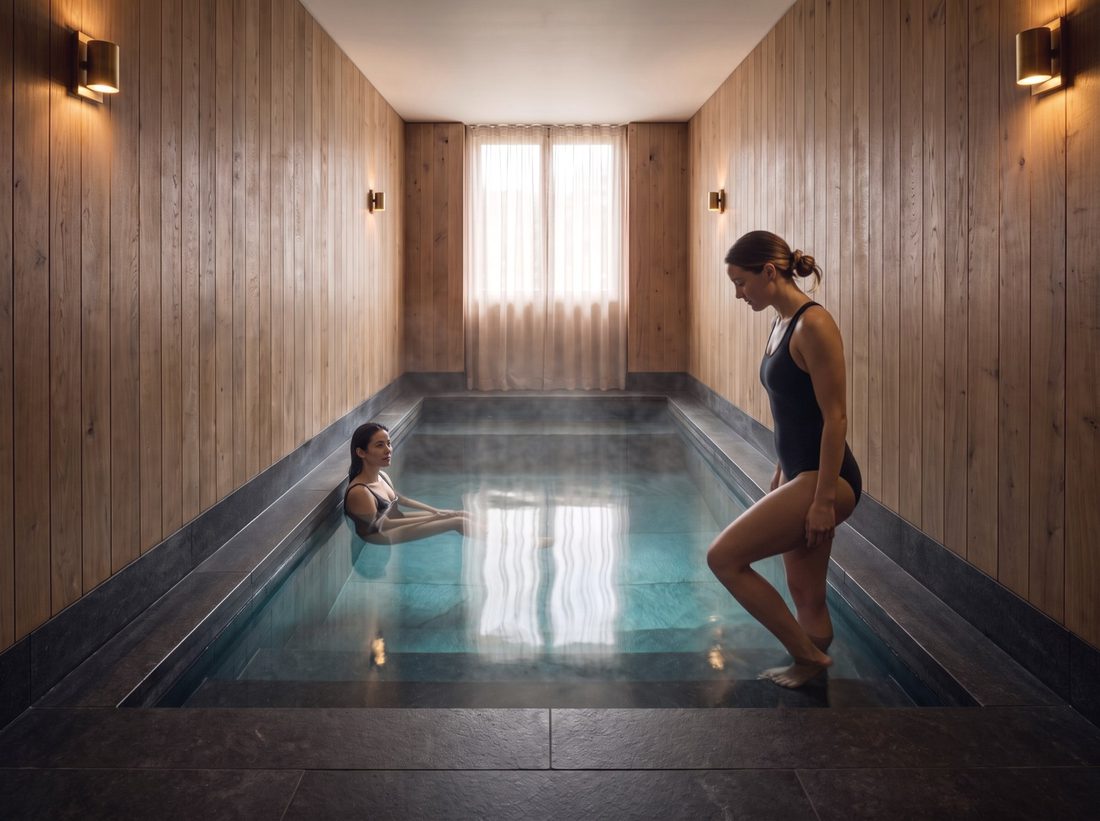 The cold tub: clear water in dark stone, warm oak walls and brass sconces, one guest resting in the water.