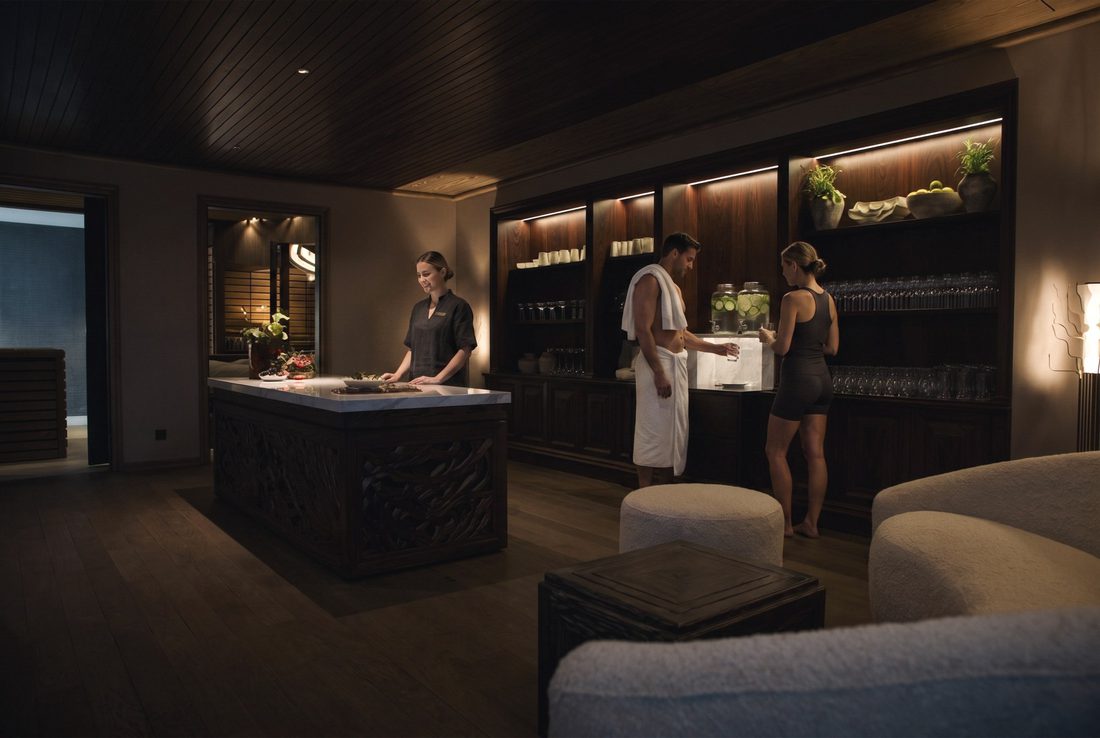 Reception: a marble-topped oak desk and the lit water station, guests in sauna wear, a bouclé sofa in the foreground.
